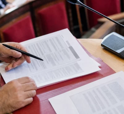 Mains anonymes feuilletant un dossier parlementaire près d’un micro de commission à l’Assemblée nationale
