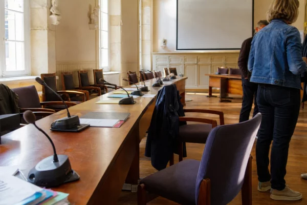Salle municipale lumineuse avec micros, dossiers et chaises vides après une réunion sur la reconstruction post-incendie.
