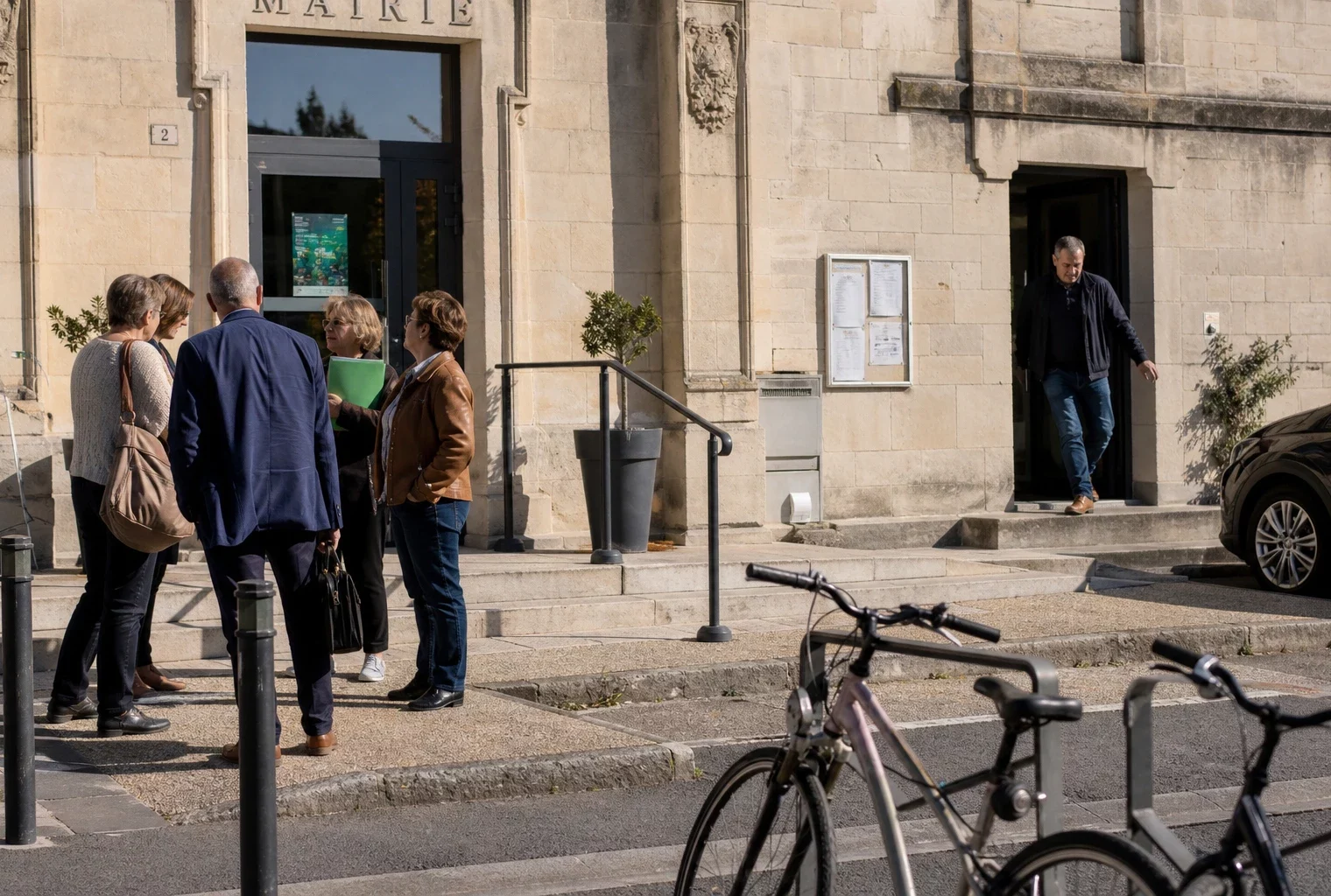 Des citoyens discutent sur le parvis d’une mairie de province, dans une scène locale claire et réaliste.