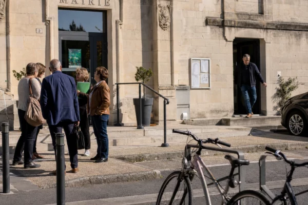 Des citoyens discutent sur le parvis d’une mairie de province, dans une scène locale claire et réaliste.