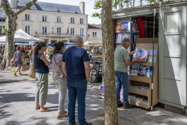 Kiosque à journaux dans une place française, avec habitants anonymes et magazines aux couvertures floutées