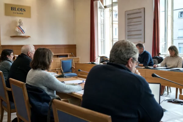Salle municipale française avec micros, dossiers flous et chaise vide lors d’un débat politique