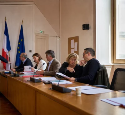 Salle municipale française avec chaise vide, micros et dossiers lors d’une réunion politique locale