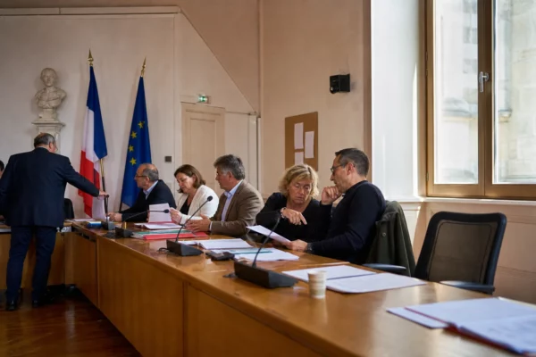 Salle municipale française avec chaise vide, micros et dossiers lors d’une réunion politique locale