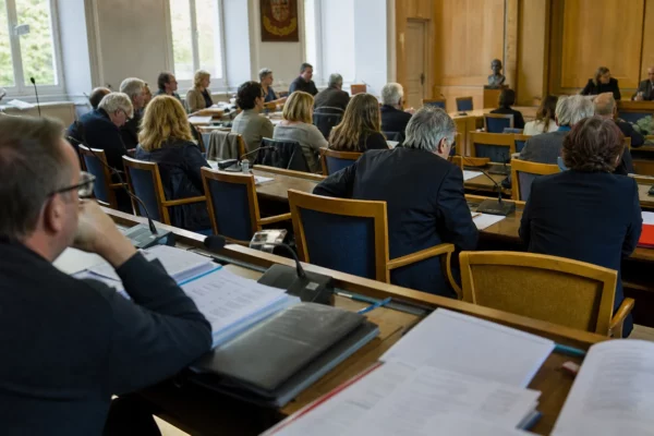 Salle municipale française avec micros, dossiers flous et une chaise vide lors d’un débat politique