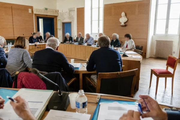 Salle de conseil municipal française avec une chaise vide, des micros et des dossiers lors d’une réunion politique