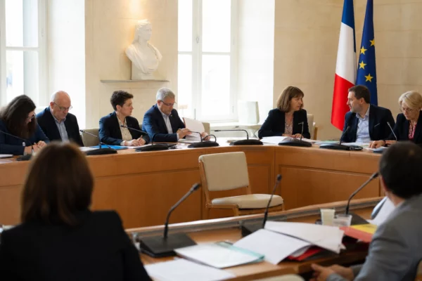 Salle municipale claire avec micros, dossiers flous et chaise vide pour illustrer la primaire socialiste.