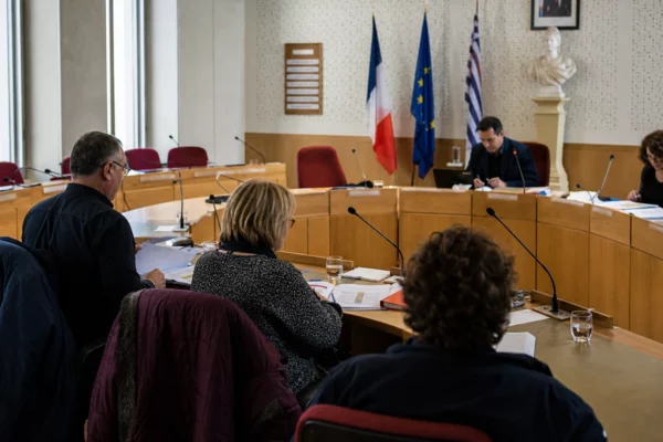 Salle municipale française préparant une primaire, avec micros, dossiers flous et une chaise vide parmi les élus