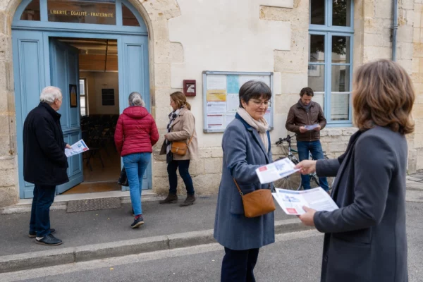 Habitants anonymes devant une mairie française avant une réunion politique locale, dans une lumière naturelle et claire.