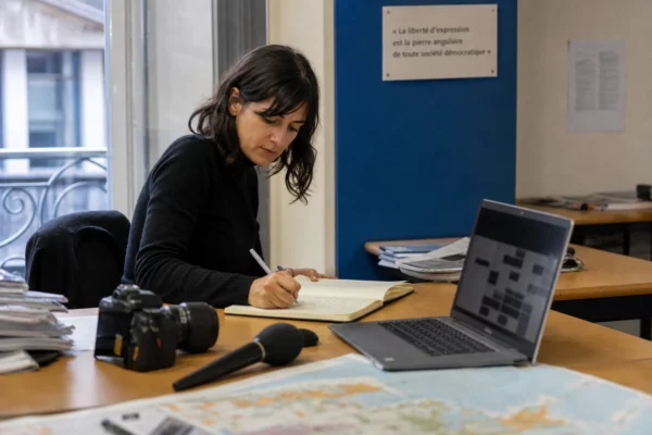 Un journaliste prépare un sujet sur Ceuta dans une rédaction lumineuse, avec carnet, micro et carte floue sur le bureau.