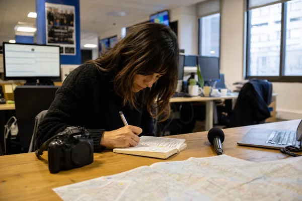 Journaliste dans une rédaction française, carnet de notes, micro sans logo et carte papier floue sur un bureau lumineux.