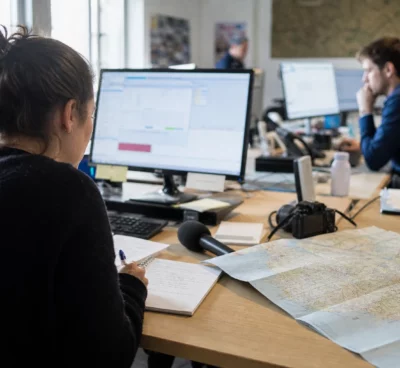 Journaliste préparant un sujet sur le budget 2027 dans une rédaction locale, avec carte régionale et micro de presse