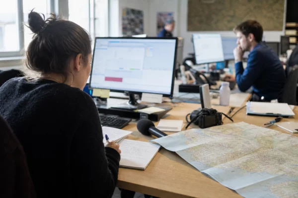 Journaliste préparant un sujet sur le budget 2027 dans une rédaction locale, avec carte régionale et micro de presse