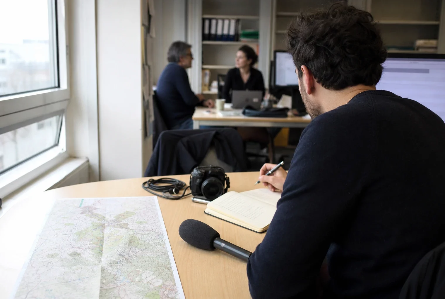 Journaliste préparant un sujet politique dans une rédaction locale française, avec carnet, carte floue et micro sans logo