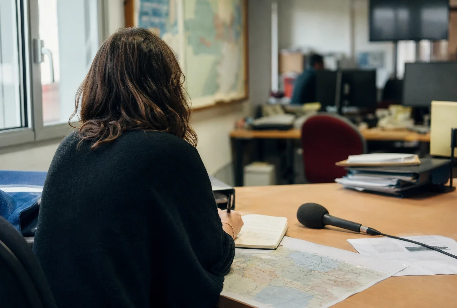 Journaliste préparant un sujet dans une rédaction locale française, avec carnet, micro et carte floue sur le bureau