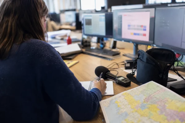 Journaliste préparant un sujet politique dans une rédaction locale française, avec carnet, micro et carte régionale floue