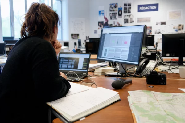 Journaliste dans une rédaction locale préparant un sujet territorial avec carnet, carte floue et micro sans logo.
