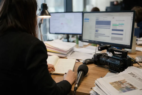 Dans une rédaction, un journaliste prépare un sujet politique avec micro, carnet et écrans flous sous une lumière naturelle.