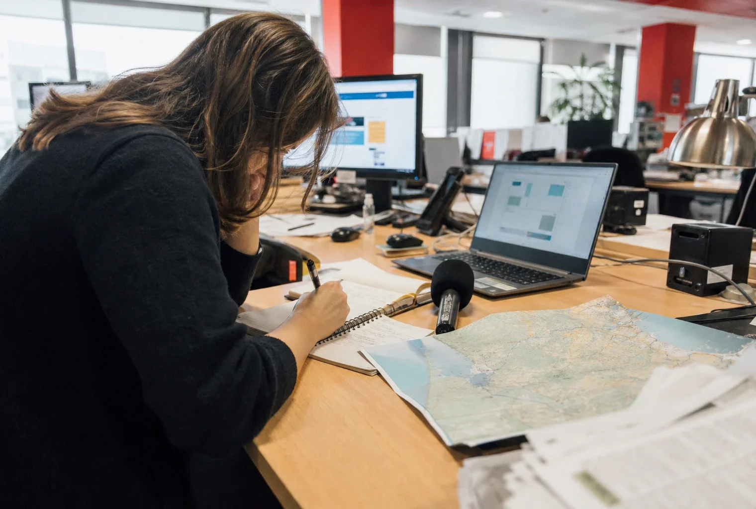 Journaliste en rédaction préparant un sujet sur la reconstruction en Gironde, avec carnet, micro et écran flou.