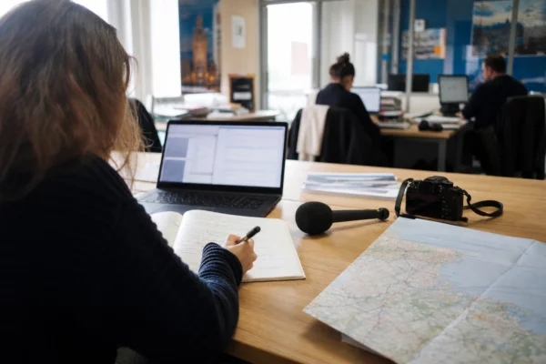 Journaliste préparant un sujet politique territorial dans une rédaction locale française, avec carnet et carte floue