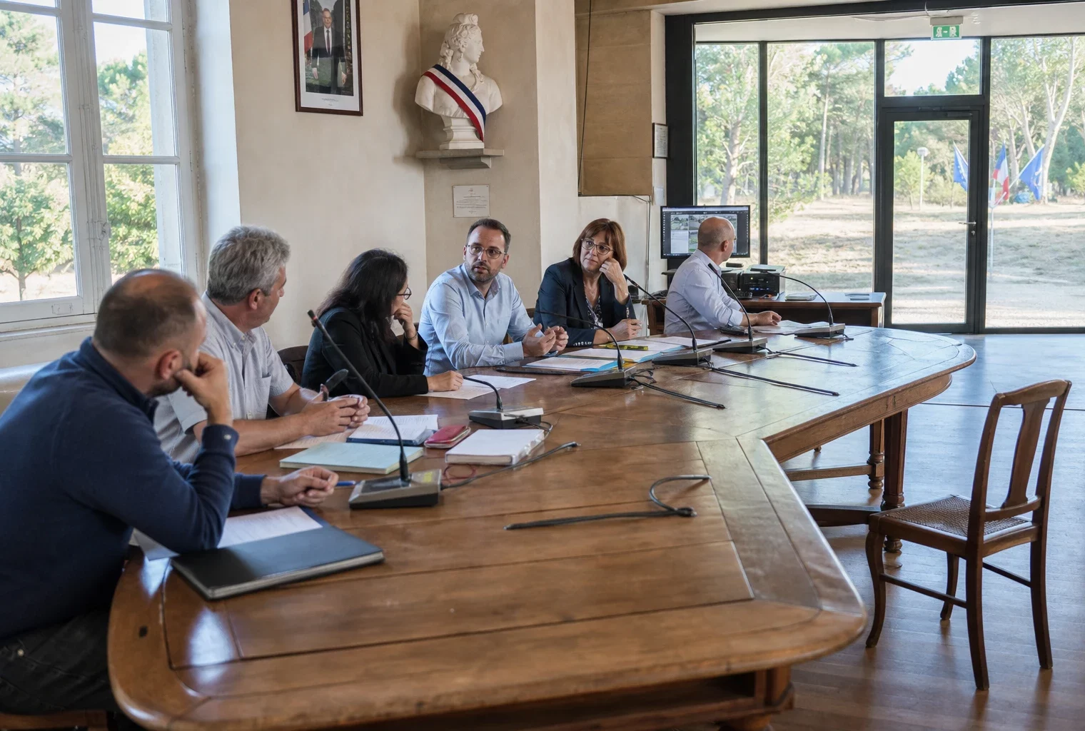 Salle municipale française avec élus anonymes, chaise vide, micros et dossiers autour d’une table de conseil