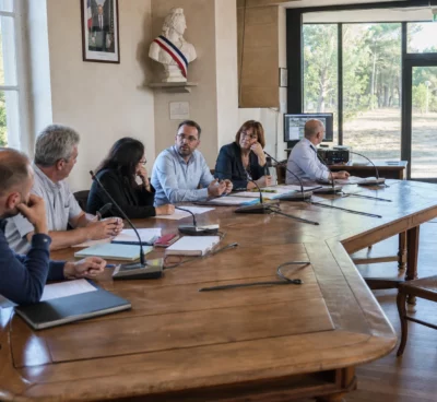 Salle municipale française avec élus anonymes, chaise vide, micros et dossiers autour d’une table de conseil