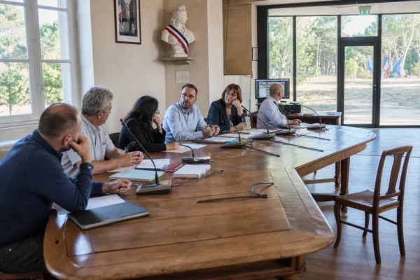 Salle municipale française avec élus anonymes, chaise vide, micros et dossiers autour d’une table de conseil