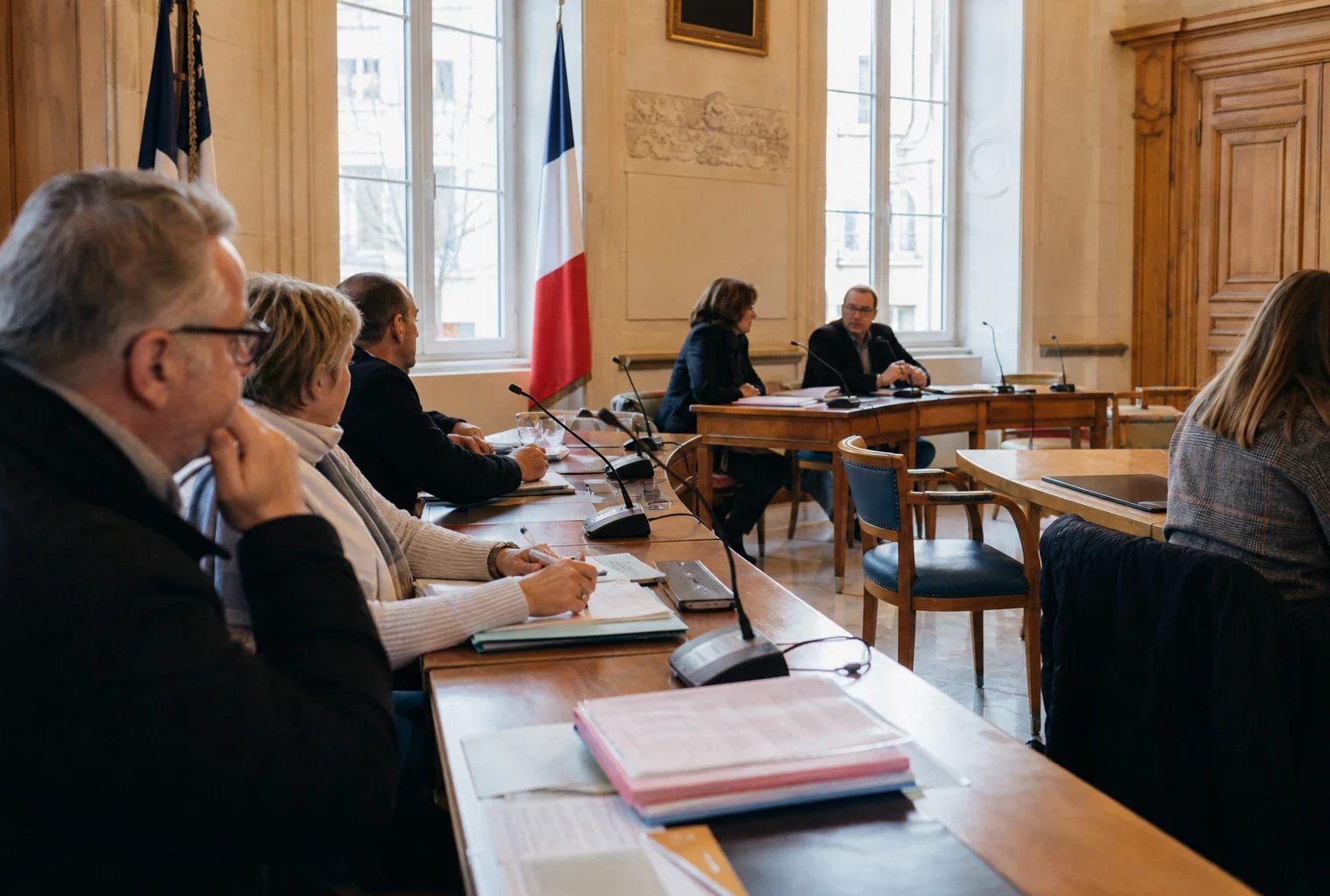 Salle municipale lumineuse avec micros, dossiers flous et chaise vide lors d’un débat local sur les incendies.