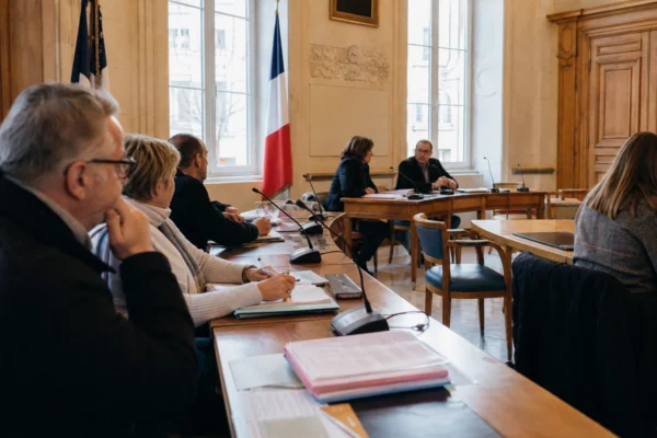 Salle municipale lumineuse avec micros, dossiers flous et chaise vide lors d’un débat local sur les incendies.