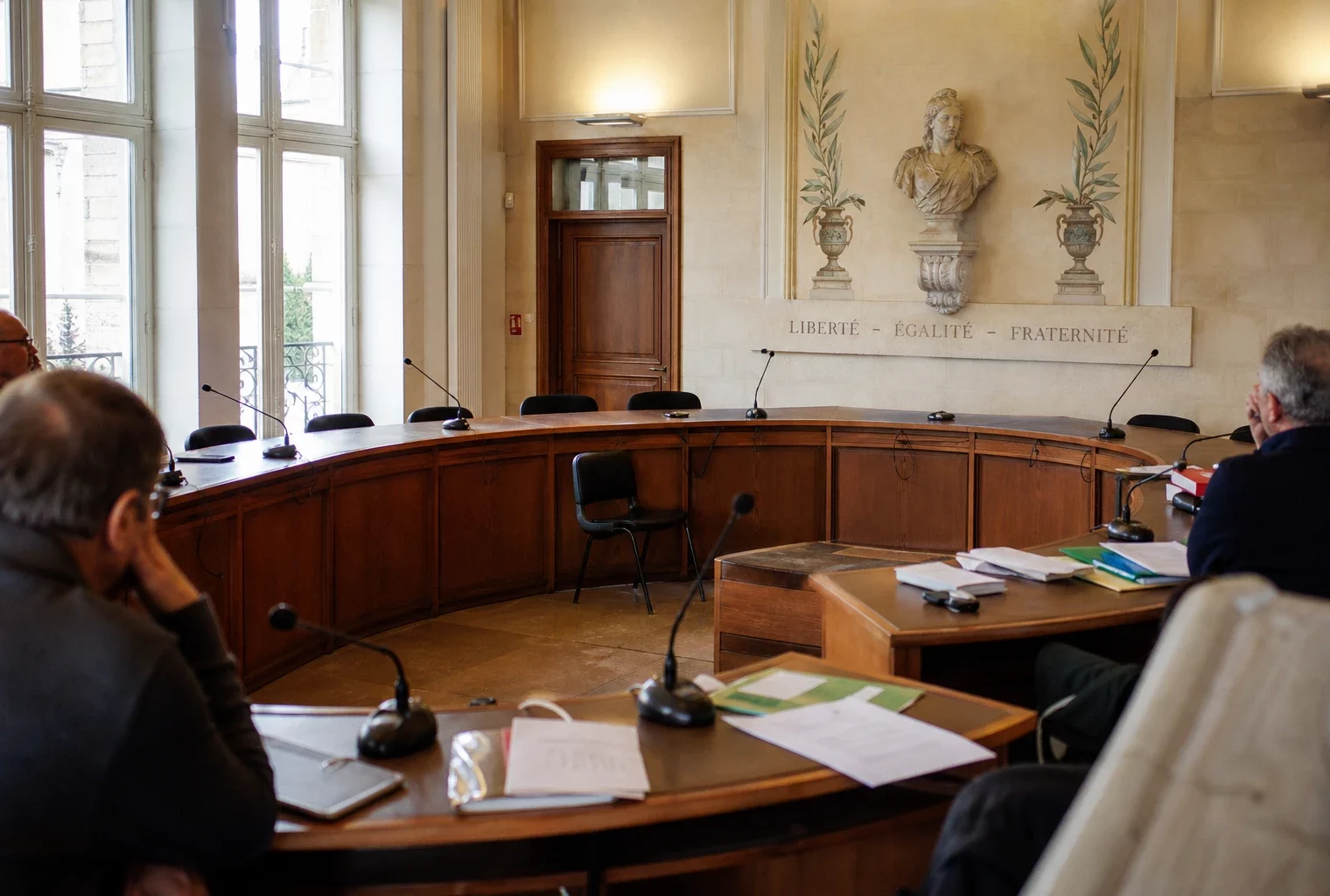 Salle municipale lumineuse avec micros, dossiers flous et chaise vide lors d’un conseil local