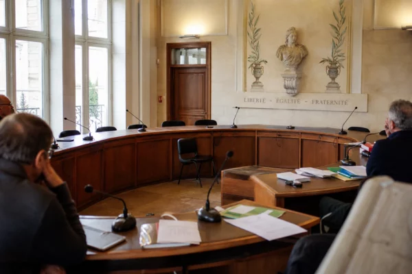 Salle municipale lumineuse avec micros, dossiers flous et chaise vide lors d’un conseil local