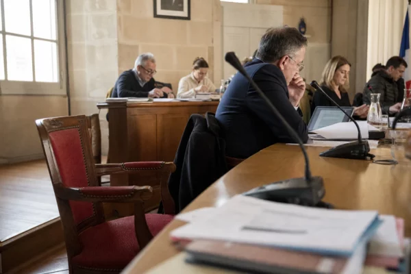 Salle municipale en plan large avec chaise vide, micros et dossiers flous lors d’un conseil local.