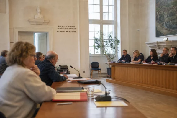 Réunion municipale dans une salle claire avec micros, dossiers flous et chaise vide subtile