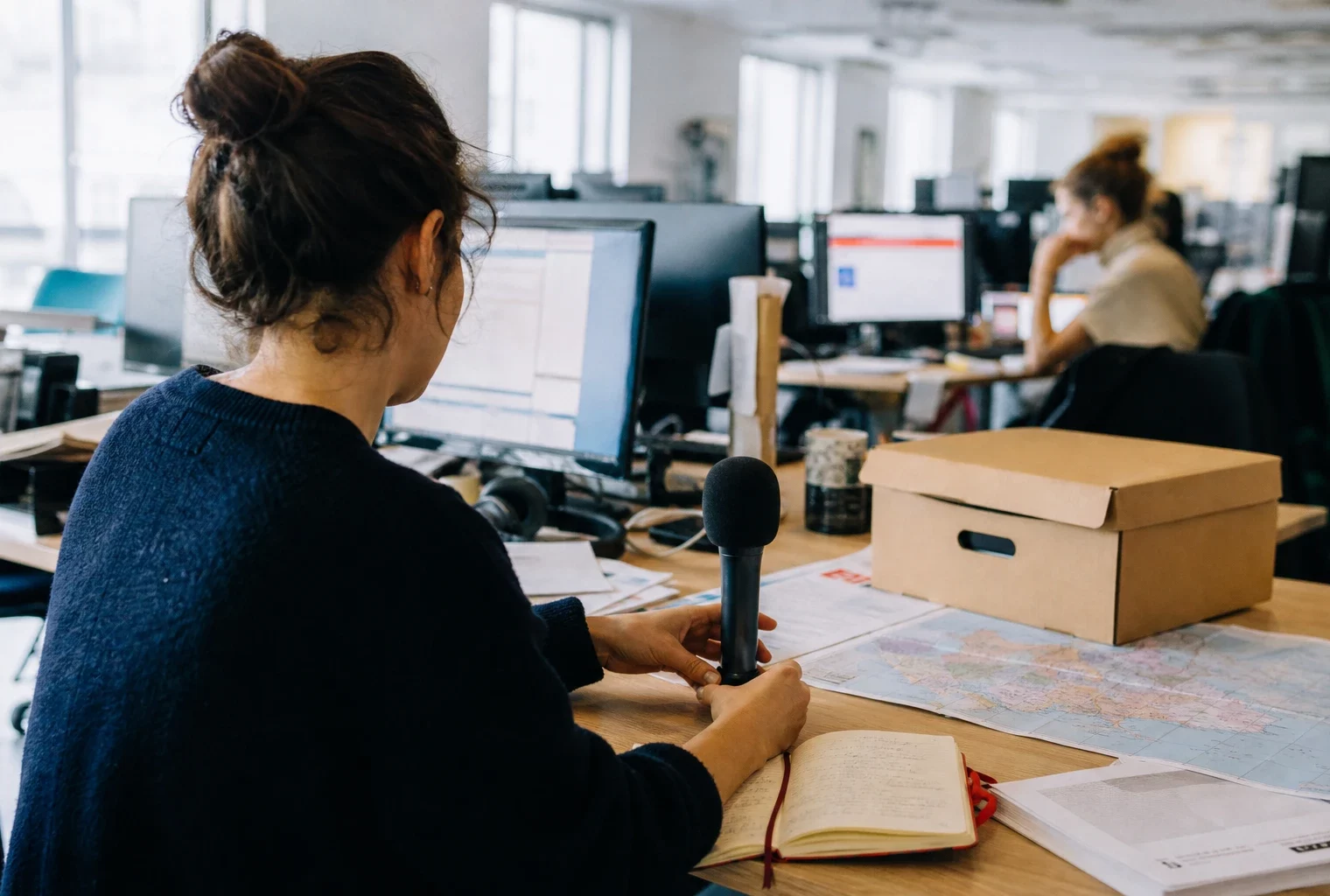 Journaliste préparant un sujet politique dans une rédaction française, avec carnet, micro et carton d’archives flou