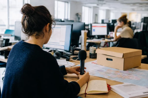 Journaliste préparant un sujet politique dans une rédaction française, avec carnet, micro et carton d’archives flou