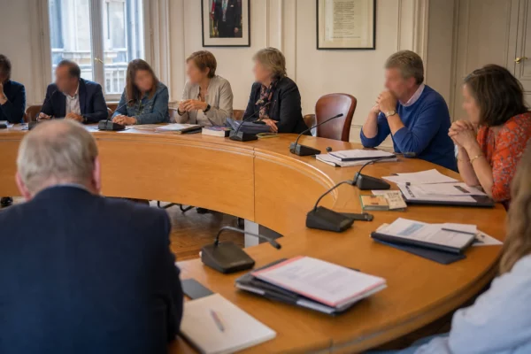 Salle municipale française avec élus anonymes, micros, dossiers flous et une chaise vide autour d’une table