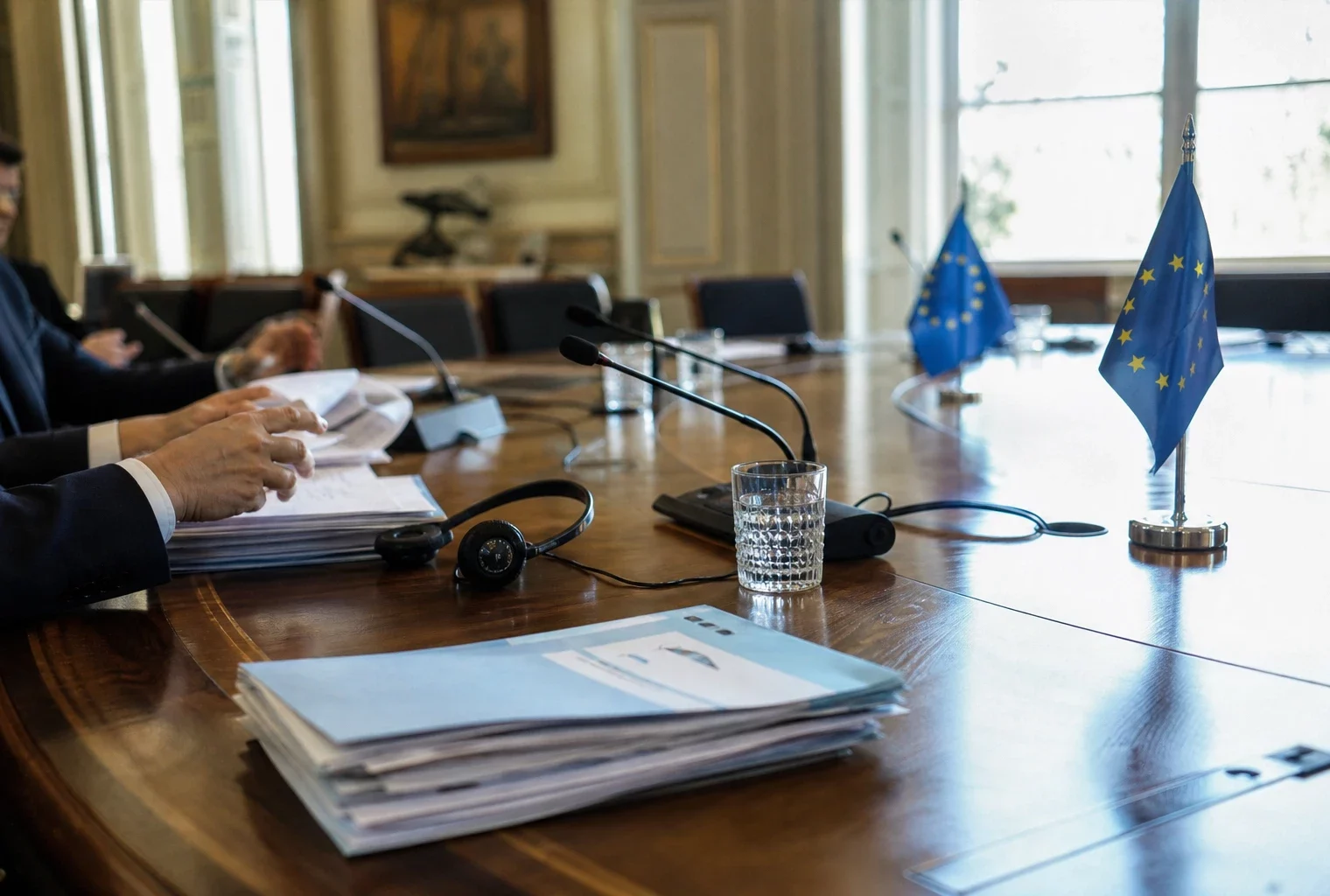 Table de négociation européenne avec dossiers, casques, verres d’eau et drapeaux miniatures, silhouettes anonymes