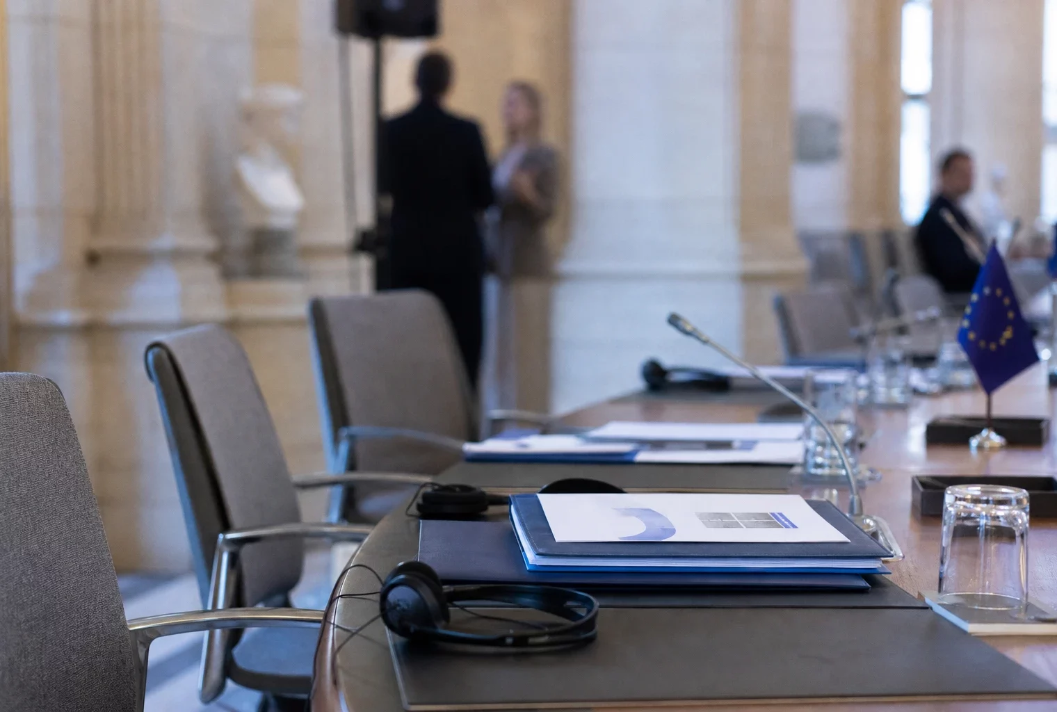 Table de négociation européenne avec casques audio, dossiers, verres d’eau et mini-drapeaux discrets, scène diplomatique réaliste.