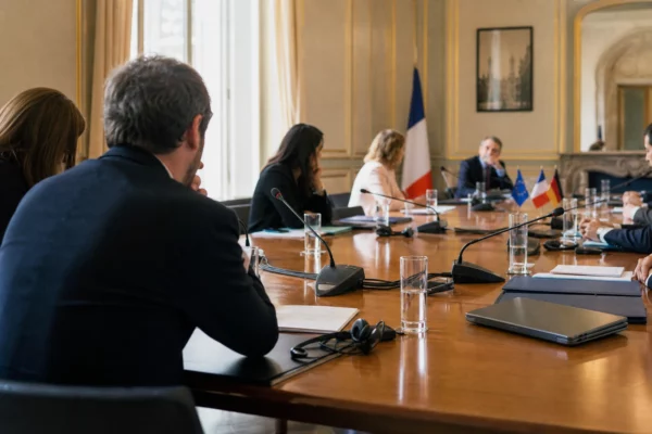 Table de négociation institutionnelle avec micros, dossiers fermés et silhouettes anonymes sous une lumière claire.