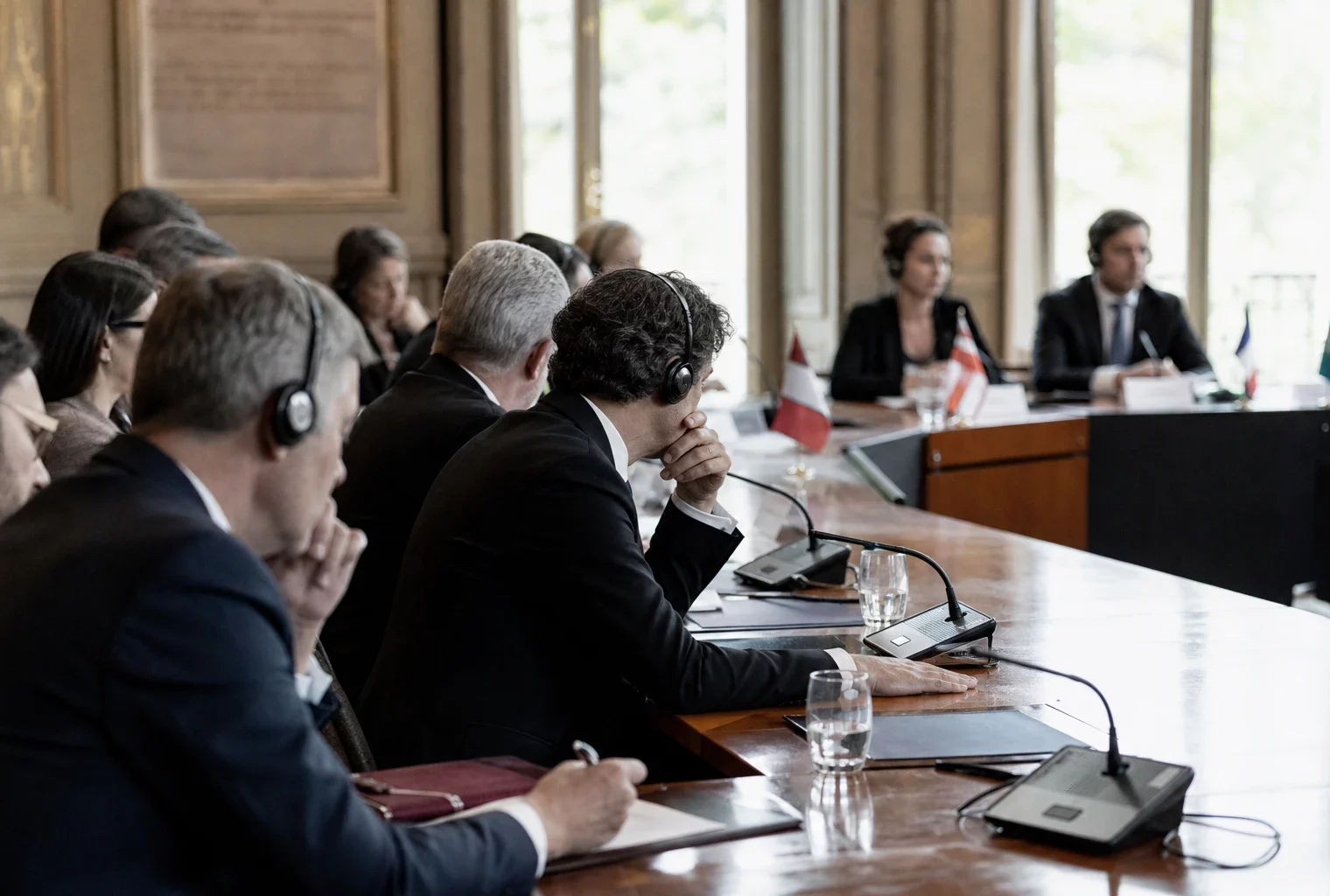 Table de négociation diplomatique avec silhouettes anonymes, micros, dossiers fermés et écouteurs dans une salle lumineuse.