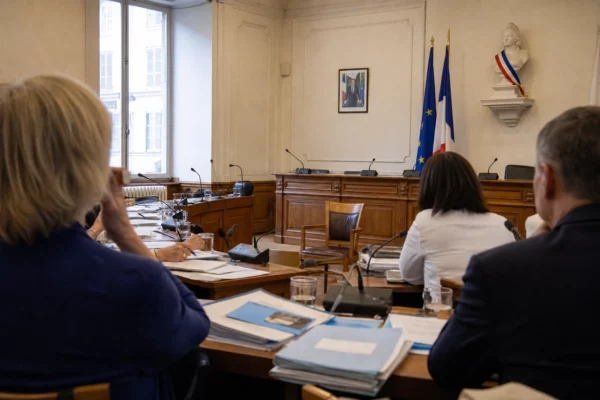 Salle municipale française avec micros, dossiers flous et une chaise vide lors d’un débat sur l’accord migratoire