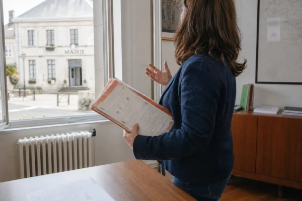 Collaborateur municipal anonymisé consultant un dossier dans une mairie française, sur fond de rue locale claire
