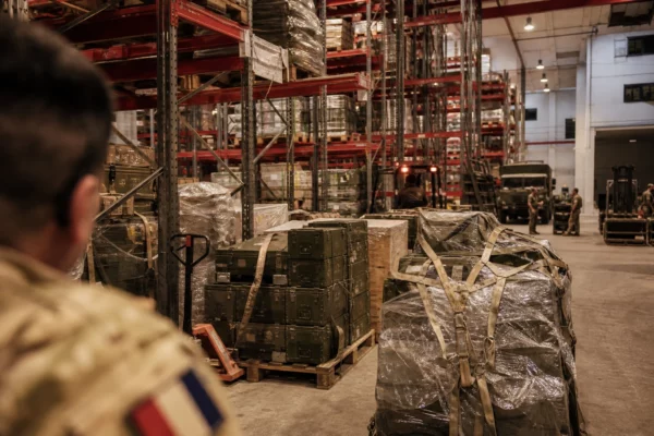 Dépôt logistique militaire en France avec palettes de caisses, sangles et chariots sous éclairage intérieur chaud.