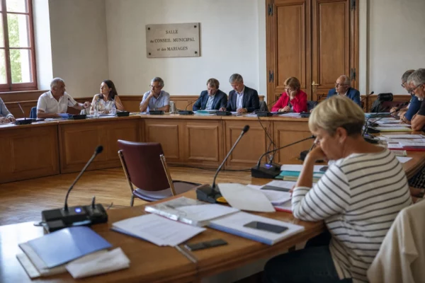 Salle municipale française avec élus anonymes, micros et dossiers autour d’une table, une chaise vide au premier plan