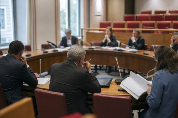 Réunion de commission parlementaire avec élus anonymes, micros et dossiers autour d’une table en lumière naturelle