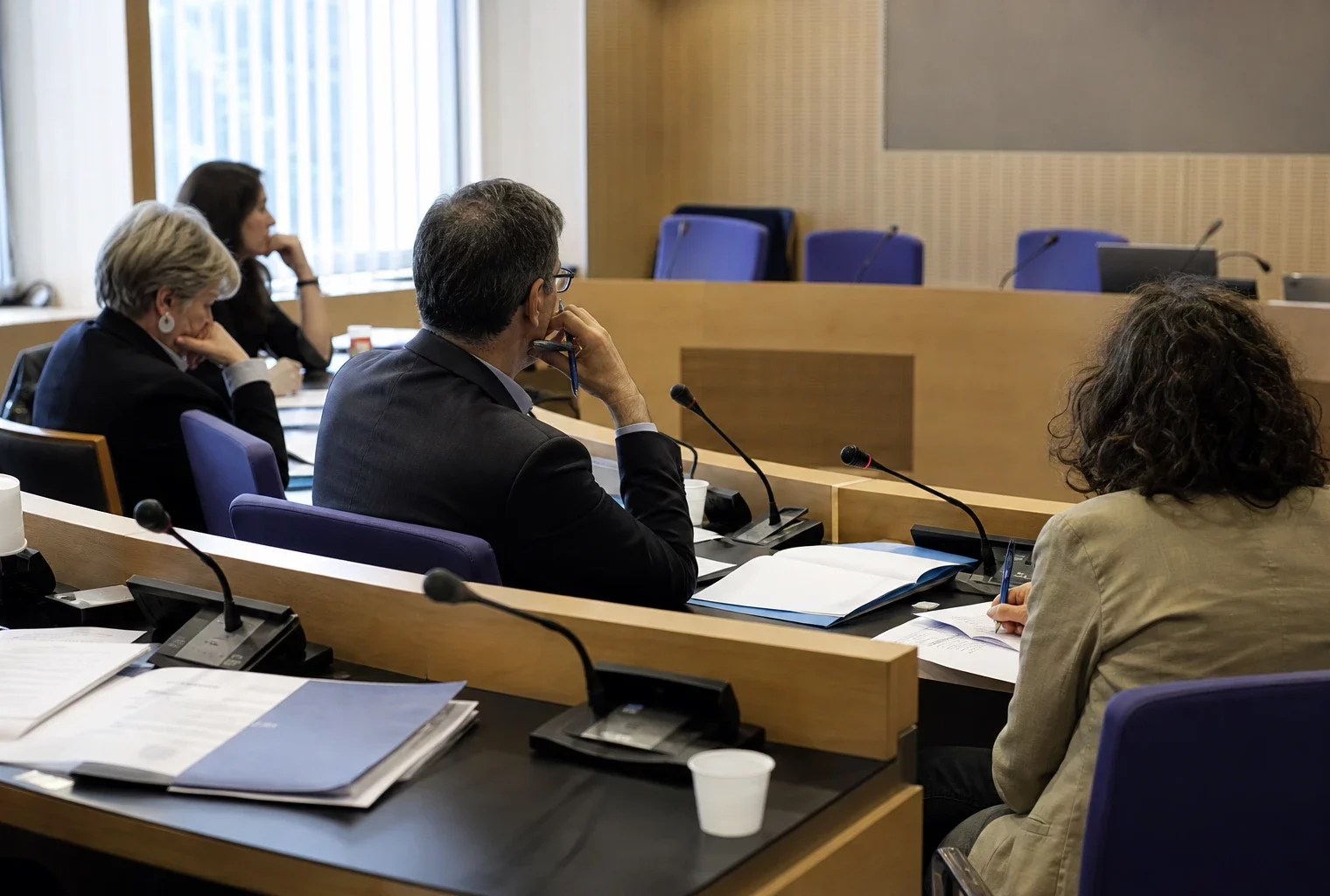 Audition parlementaire française avec silhouettes anonymes, micros et dossiers sur une table éclairée naturellement