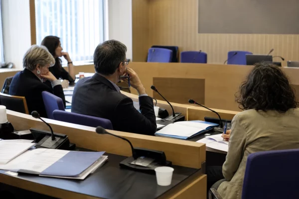 Audition parlementaire française avec silhouettes anonymes, micros et dossiers sur une table éclairée naturellement