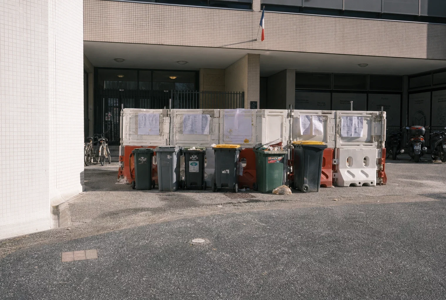 Devant un lycée bloqué, barrières, poubelles et vélos stationnés sous un soleil de fin de matinée.