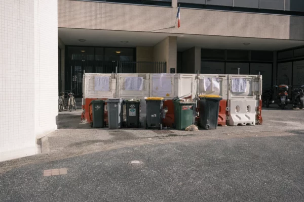 Devant un lycée bloqué, barrières, poubelles et vélos stationnés sous un soleil de fin de matinée.