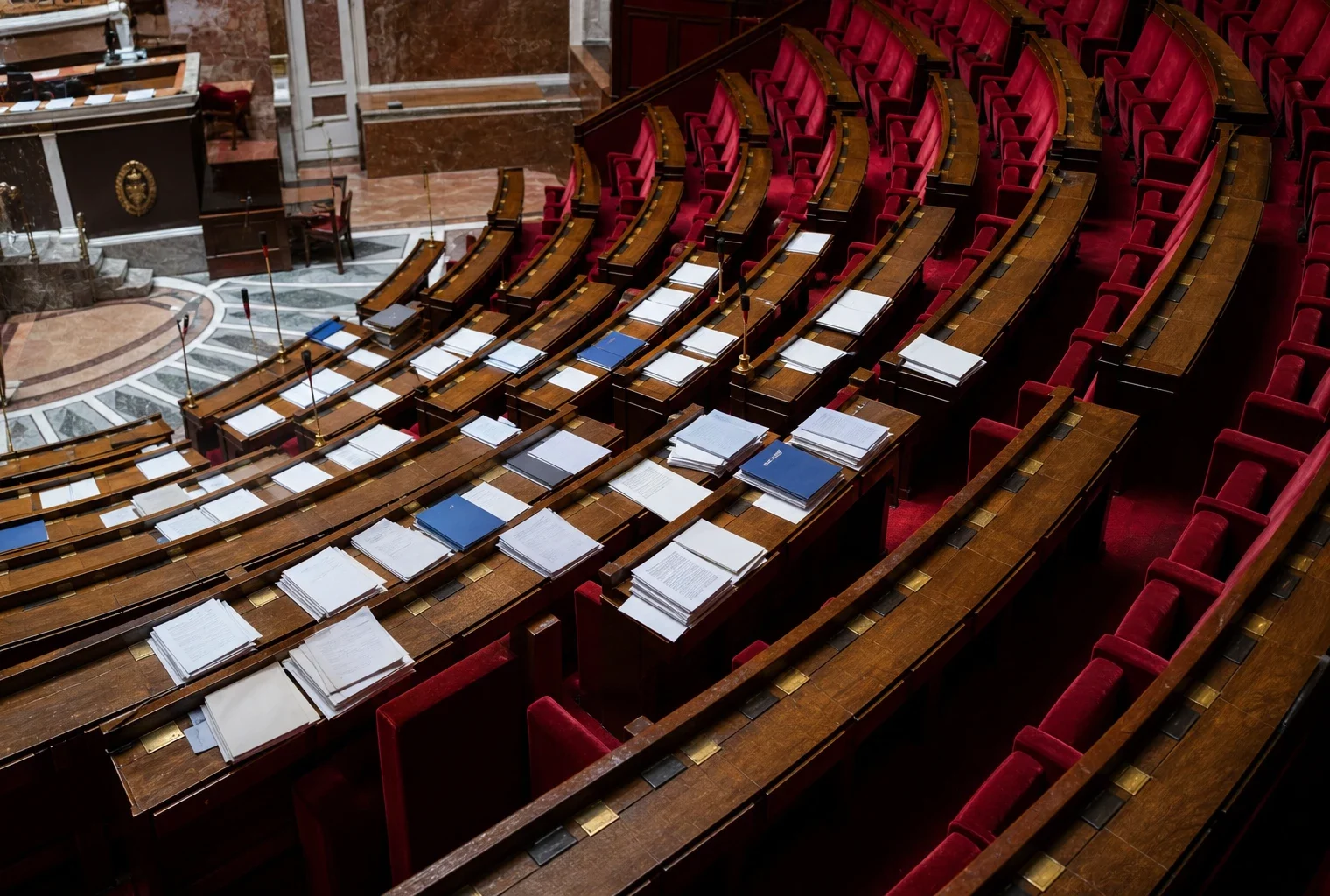 Hémicycle de l’Assemblée nationale vide, avec pupitres, micros et dossiers de séance au centre, vu en plongée.